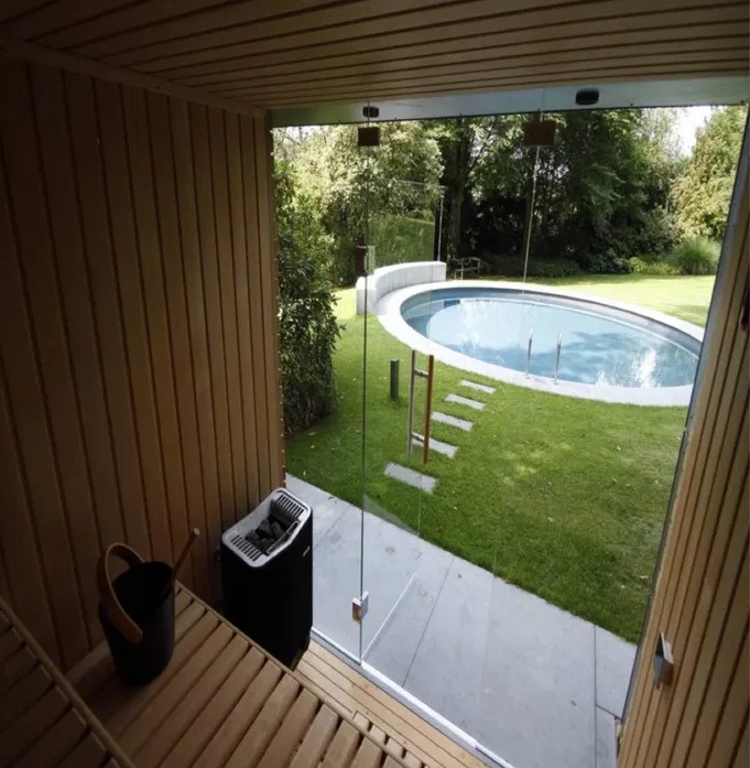 Wnętrze drewnianej sauny z widokiem na basen w ogrodzie przez szklane drzwi. Widoczny piec do sauny i wiadro z chochlą.