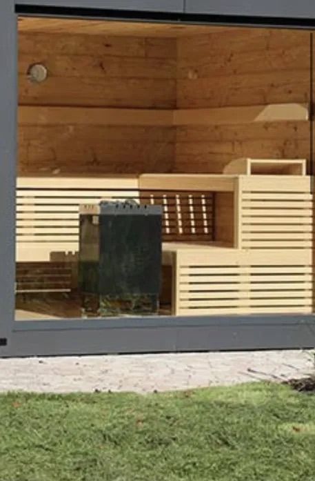 Nowoczesna sauna z jasnego drewna z piecem elektrycznym za szklaną osłoną, widoczna z zewnątrz przez szklane drzwi przesuwne na tle trawnika.