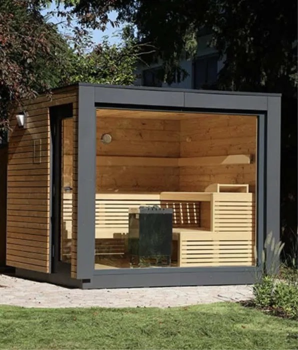 Nowoczesna sauna ogrodowa z drewnianym wnętrzem i zewnętrzną obudową w odcieniach szarości, widoczna przez przeszkloną ścianę, otoczona zielenią.