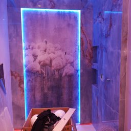 Piotr Majesz - Wykończenie łazienki z płytkami imitującymi marmur, podświetleniem LED i dekoracyjnym panelem z motywem flamingów, w trakcie prac montażowych widoczny karton z elementami wyposażenia.