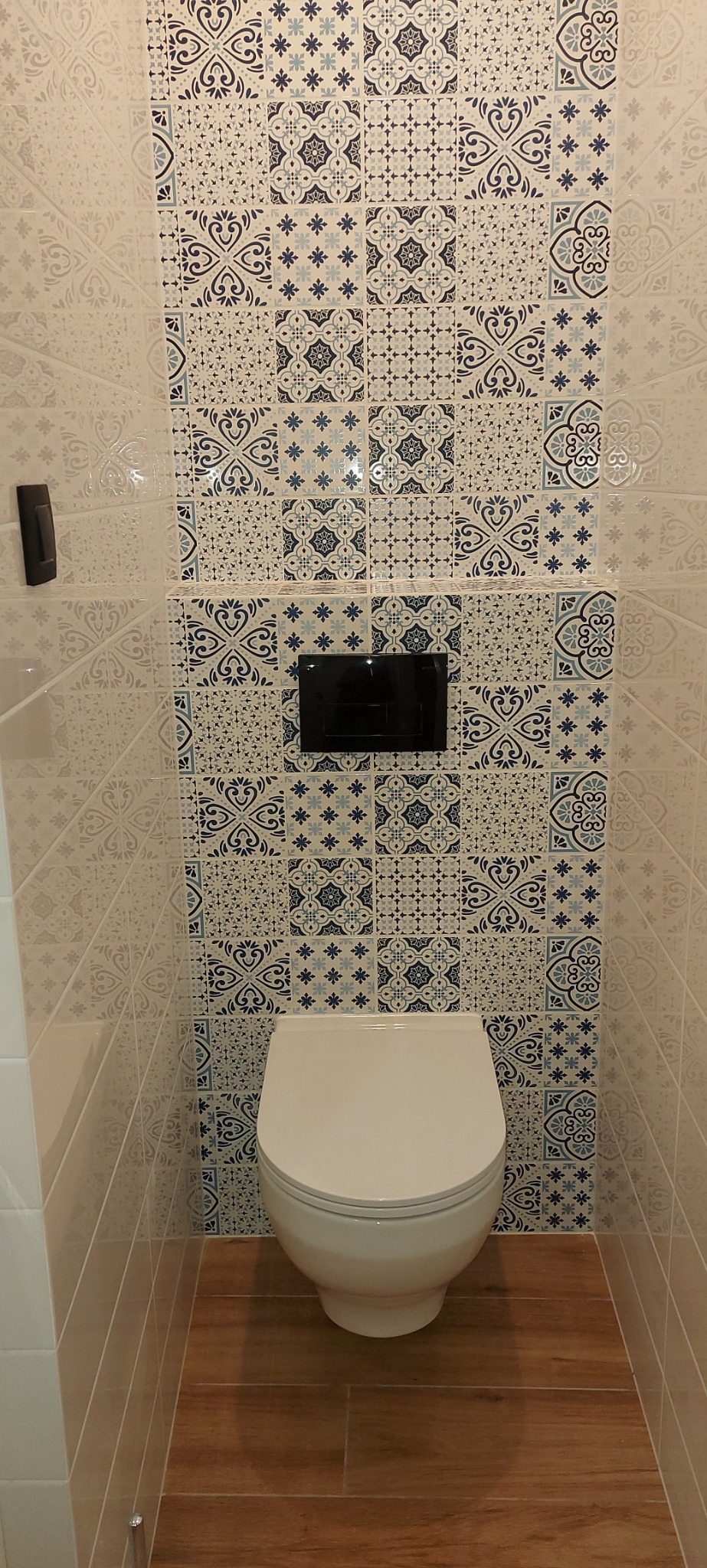 Nowoczesna toaleta z białą miską ustępową, zabudowaną spłuczką w czarnej obudowie oraz ścianą wyłożoną dekoracyjnymi płytkami w odcieniach bieli i niebieskiego, kontrastującymi z podłogą z płytek...