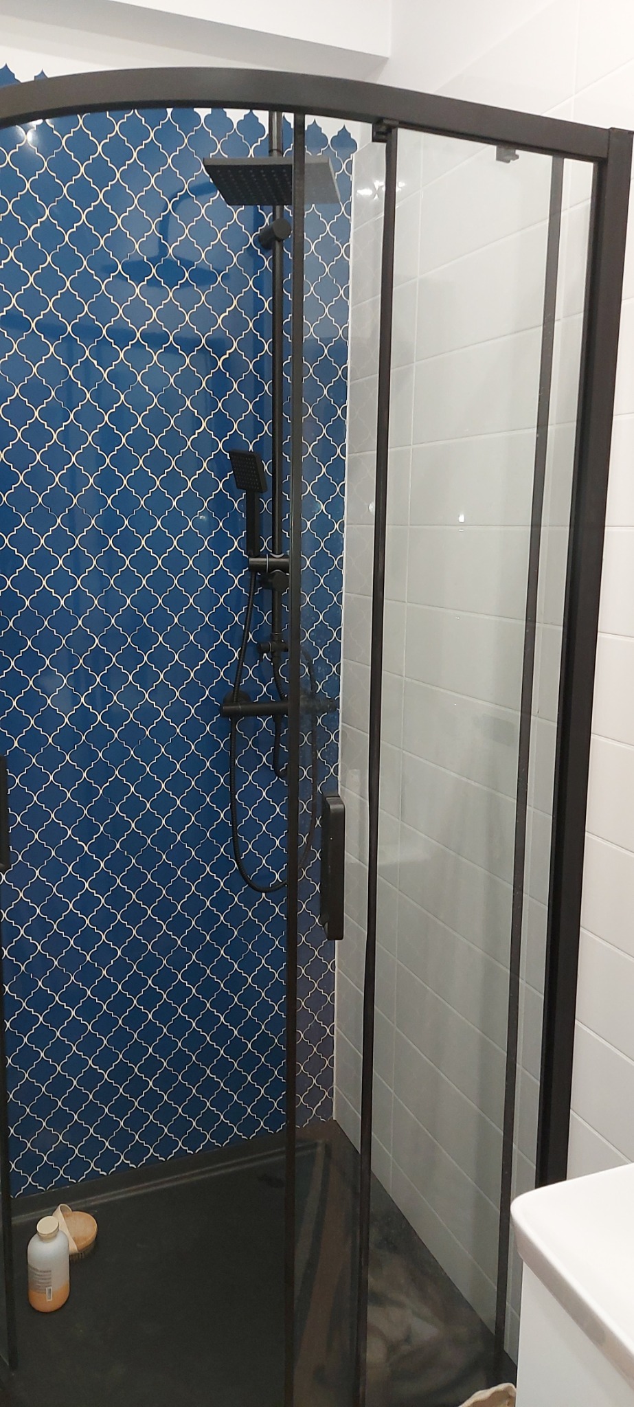 Kabina prysznicowa z czarną ramą, niebieskie płytki z białym wzorem marokańskim na ścianie, białe prostokątne płytki po prawej stronie, czarny brodzik, butelka i szczotka w rogu.