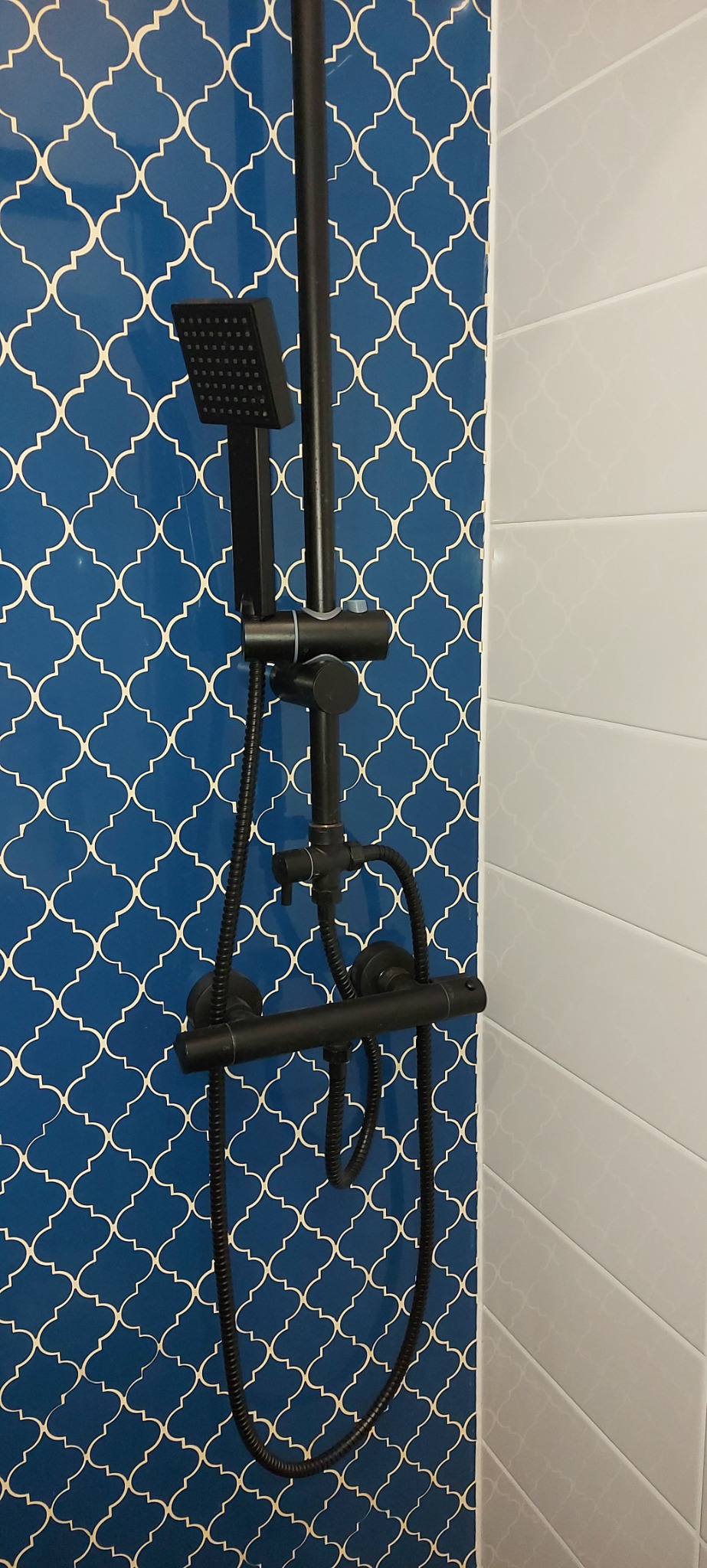 Nowoczesny, czarny zestaw prysznicowy z deszczownicą na tle ściany wyłożonej niebieskimi płytkami z marokańskim wzorem oraz białymi, poziomymi płytkami.