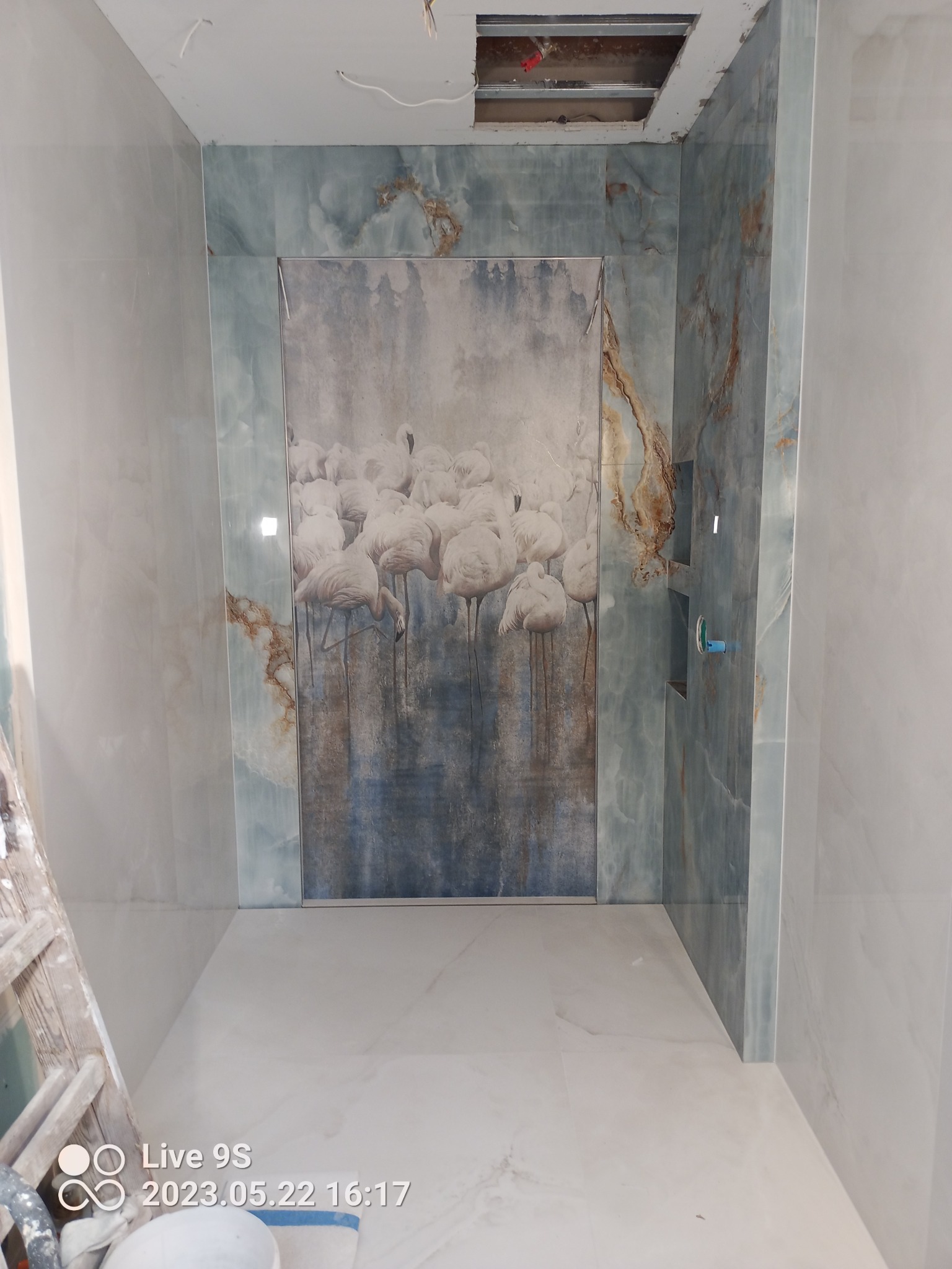 Niedokończona łazienka z płytkami imitującymi onyks w odcieniach błękitu i szarości, ozdobiona panelem z motywem flamingów. Widoczne niedokończone prace przy suficie i drabina malarska.