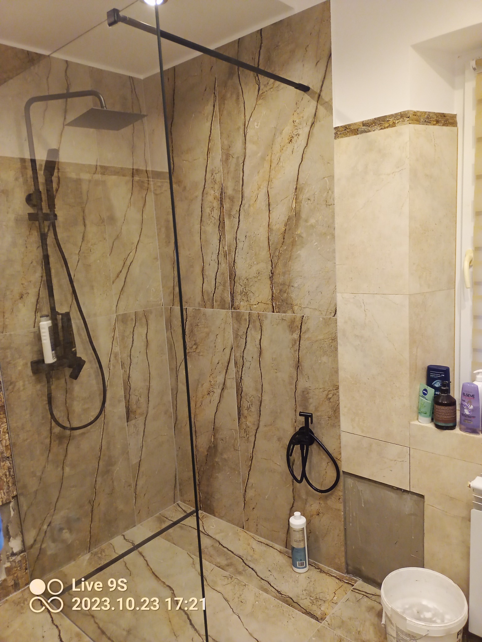 Nowoczesna kabina prysznicowa z deszczownicą i czarnymi elementami, wykończona płytkami imitującymi marmur, widoczny fragment niedokończonej zabudowy i wiadro po farbie.