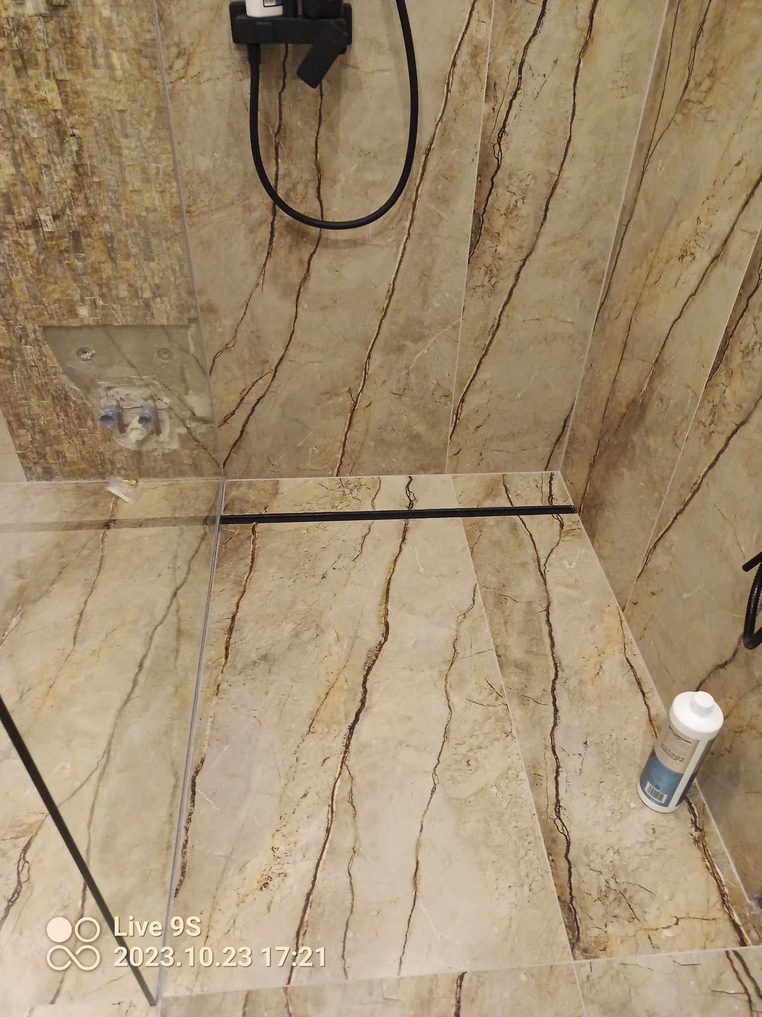 Płytki imitujące marmur w kabinie prysznicowej z widocznym odpływem liniowym i niezabudowanymi przyłączami wodnymi, butelka środka czyszczącego w rogu.