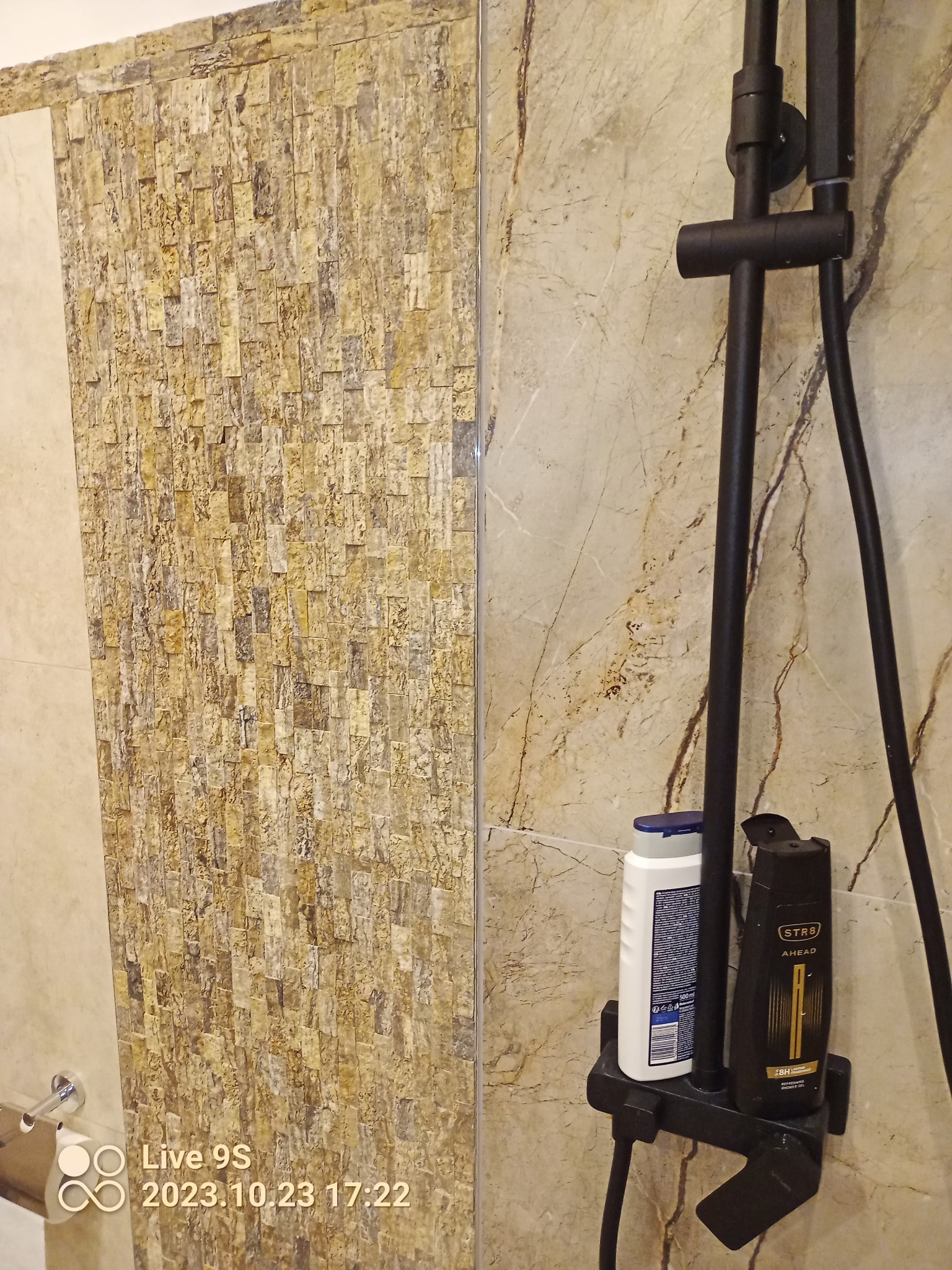 Wykończona ściana w łazience z mozaiką kamienną, widoczny czarny zestaw prysznicowy z półką na kosmetyki.