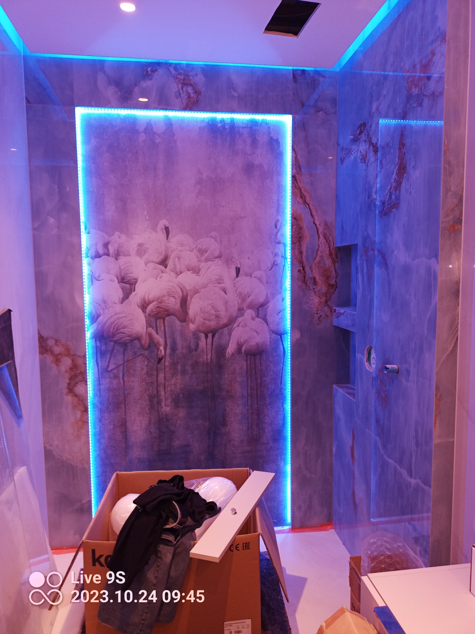 Wykończenie łazienki z płytkami imitującymi marmur, podświetleniem LED i dekoracyjnym panelem z motywem flamingów, w trakcie prac montażowych widoczny karton z elementami wyposażenia.