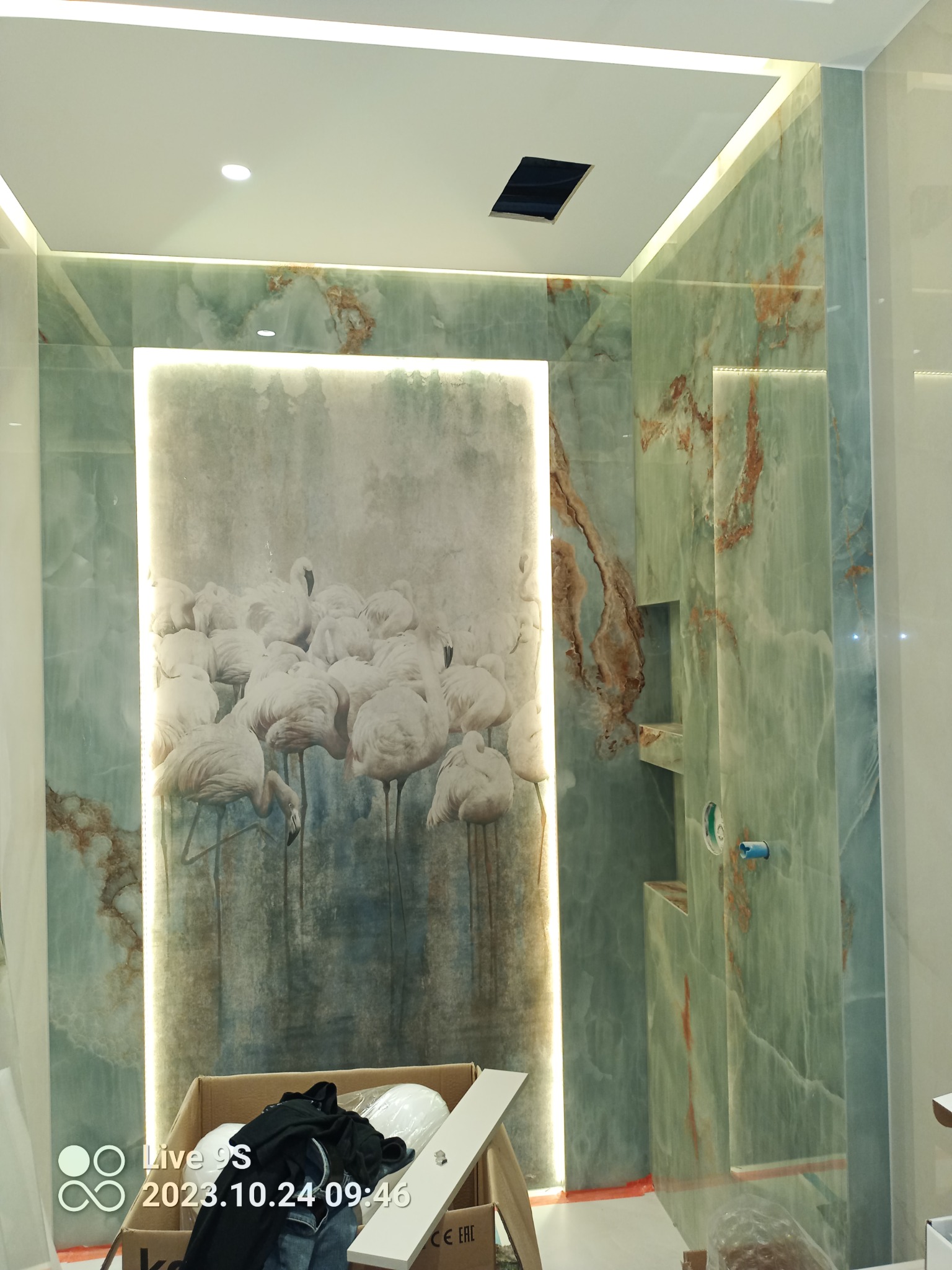 Wykończona łazienka z zielonego marmuru z podświetlanym panelem dekoracyjnym z motywem flamingów, widoczne wnęki w ścianie oraz instalacja prysznicowa, w tle karton z ubraniami.