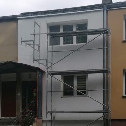 Firma Ogólnobudowlana - Fragment elewacji budynku w trakcie malowania na biało, widoczne rusztowanie, dwa kolory elewacji sąsiadują ze sobą, na dole widoczne wiaderka i elementy rusztowania.