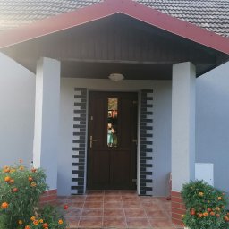 Firma Ogólnobudowlana - Ocieplony dom z szarą elewacją, brązowymi drzwiami wejściowymi i dekoracyjnymi elementami z szarej cegły, pomarańczowe nagietki przy wejściu, widoczny fragment dachu z czerwonymi elementami.