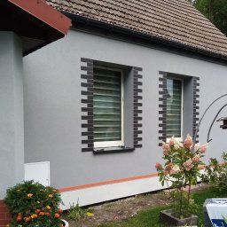 Firma Ogólnobudowlana - Szara elewacja domu jednorodzinnego z dekoracyjnym obramowaniem okien z ciemnej cegły klinkierowej, widoczne kwiaty w donicach i krzewach przed budynkiem.