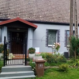 Firma Ogólnobudowlana - Szary dom jednorodzinny z brązowym dachem, czarną bramą i elementami dekoracyjnymi z cegły wokół okien, częściowo zasłonięty rusztowaniem i bujną roślinnością w ogrodzie.