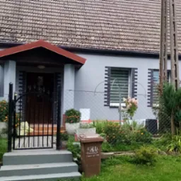 Szary dom jednorodzinny z brązowym dachem, czarną bramą i elementami dekoracyjnymi z cegły wokół okien, częściowo zasłonięty rusztowaniem i bujną roślinnością w ogrodzie.