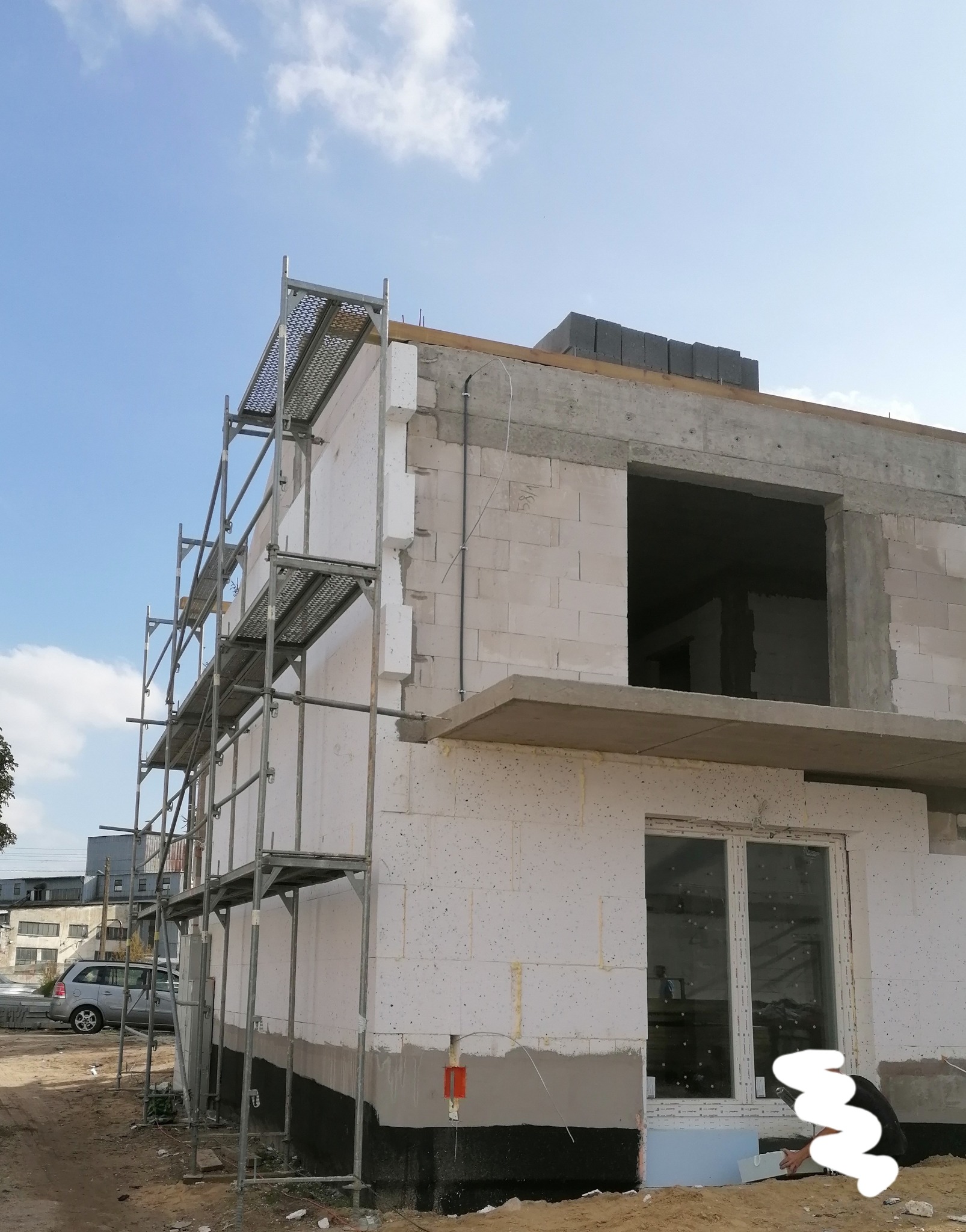 Budowa domu jednorodzinnego w trakcie ocieplania elewacji, widoczne rusztowanie, białe bloczki, fragment okna i izolacja termiczna.