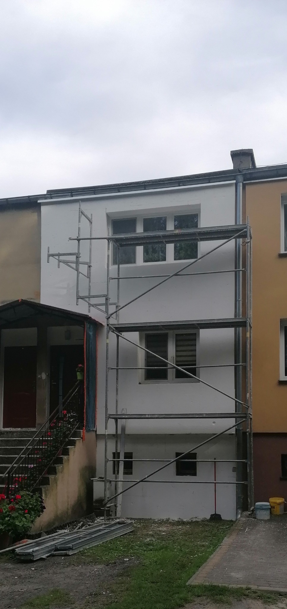 Fragment elewacji budynku w trakcie malowania na biało, widoczne rusztowanie, dwa kolory elewacji sąsiadują ze sobą, na dole widoczne wiaderka i elementy rusztowania.