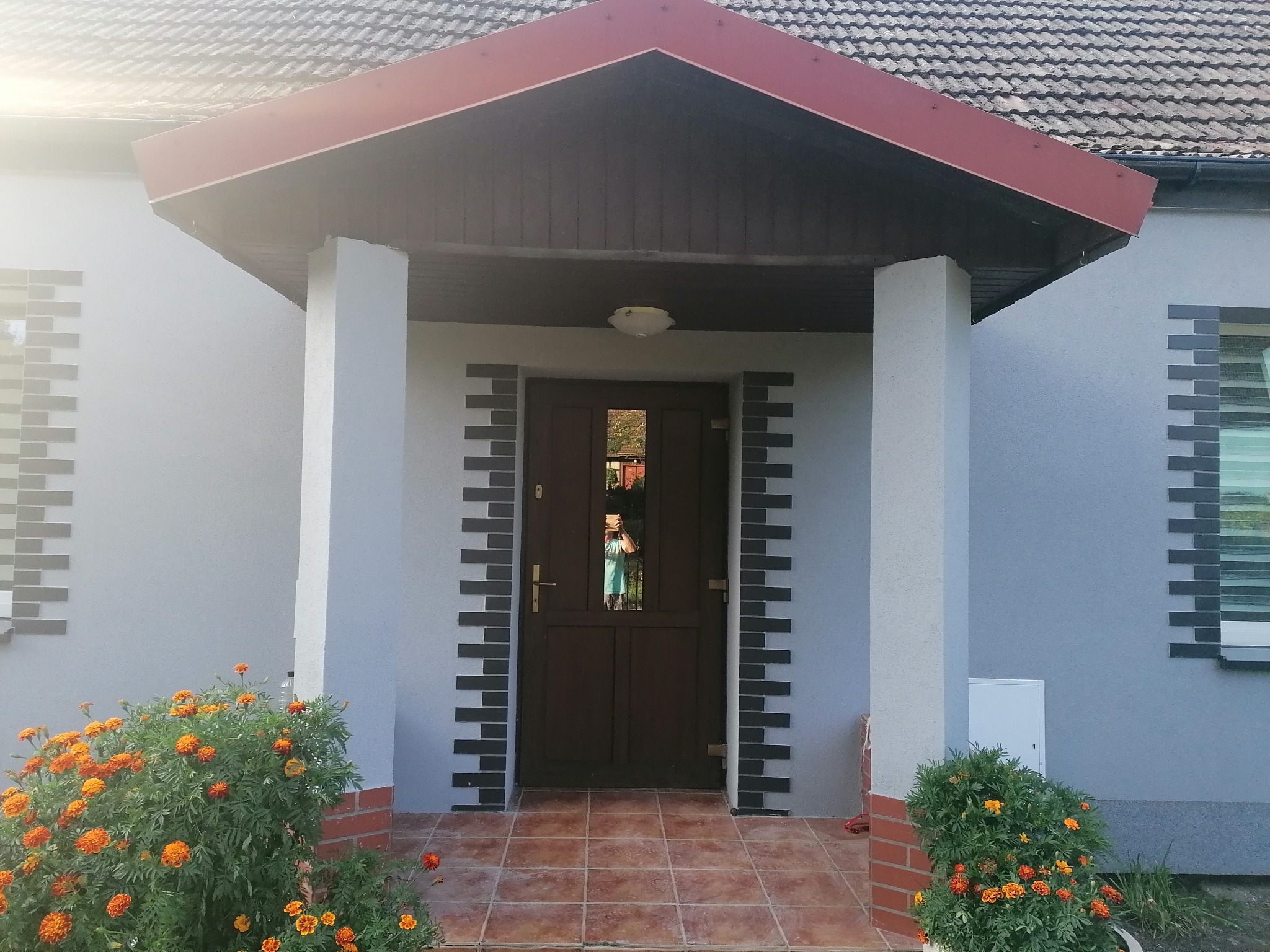 Ocieplony dom z szarą elewacją, brązowymi drzwiami wejściowymi i dekoracyjnymi elementami z szarej cegły, pomarańczowe nagietki przy wejściu, widoczny fragment dachu z czerwonymi elementami.