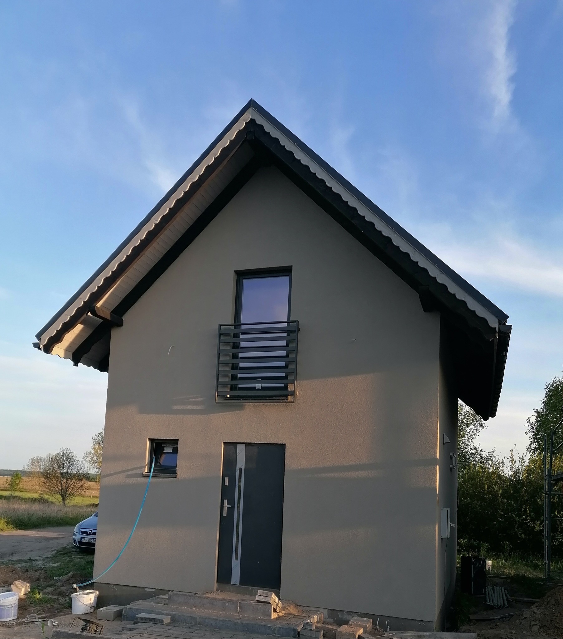 Otynkowany, szary dom jednorodzinny z czarną stolarką okienną i drzwiową, widoczny w świetle zachodzącego słońca, z niebieskim wężem ogrodowym wystającym z okna na parterze.