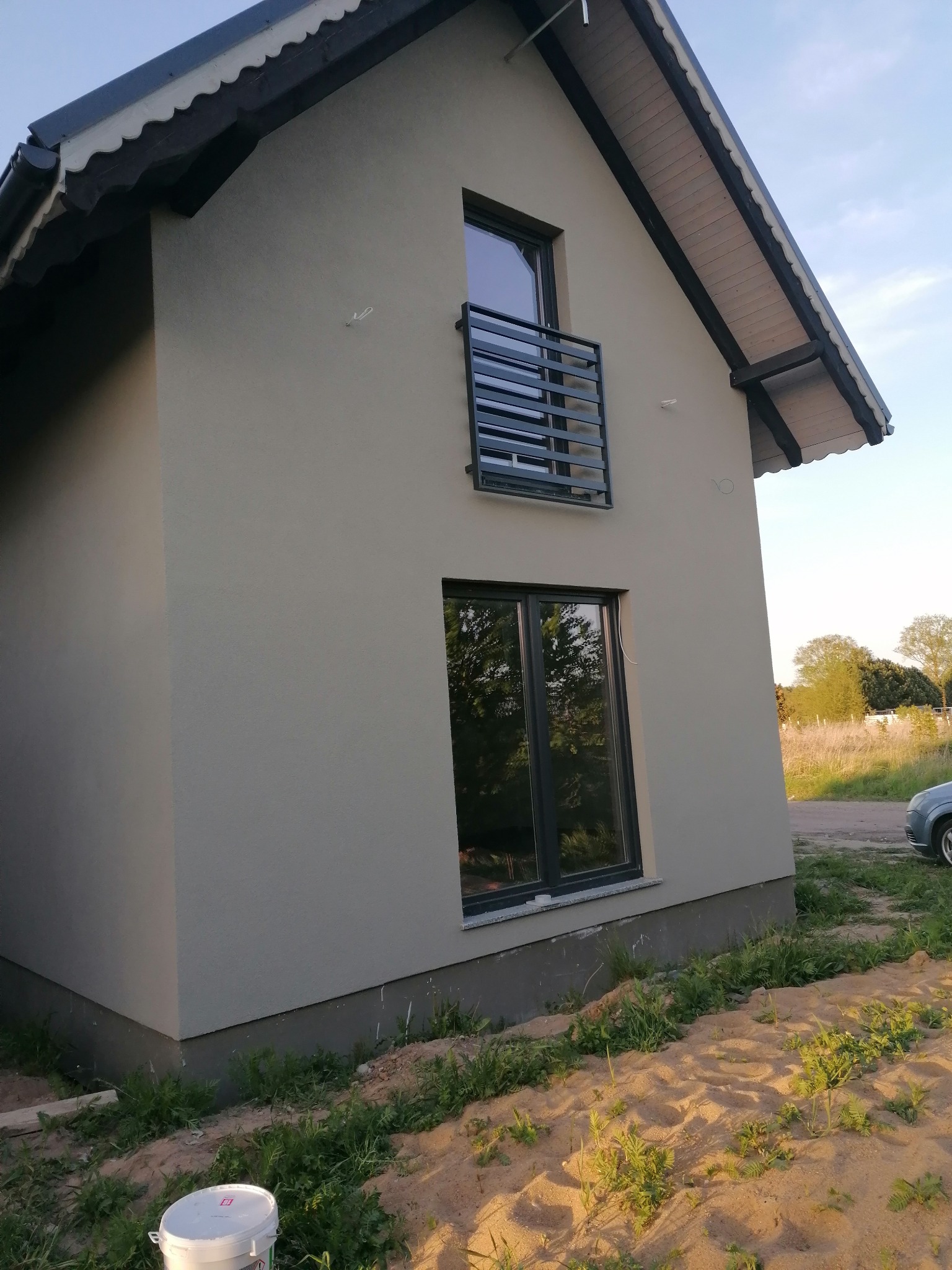 Szary dom jednorodzinny po ociepleniu elewacji, widoczny z boku, z balkonem i oknami z czarnymi ramami, na pierwszym planie wiadro po farbie.