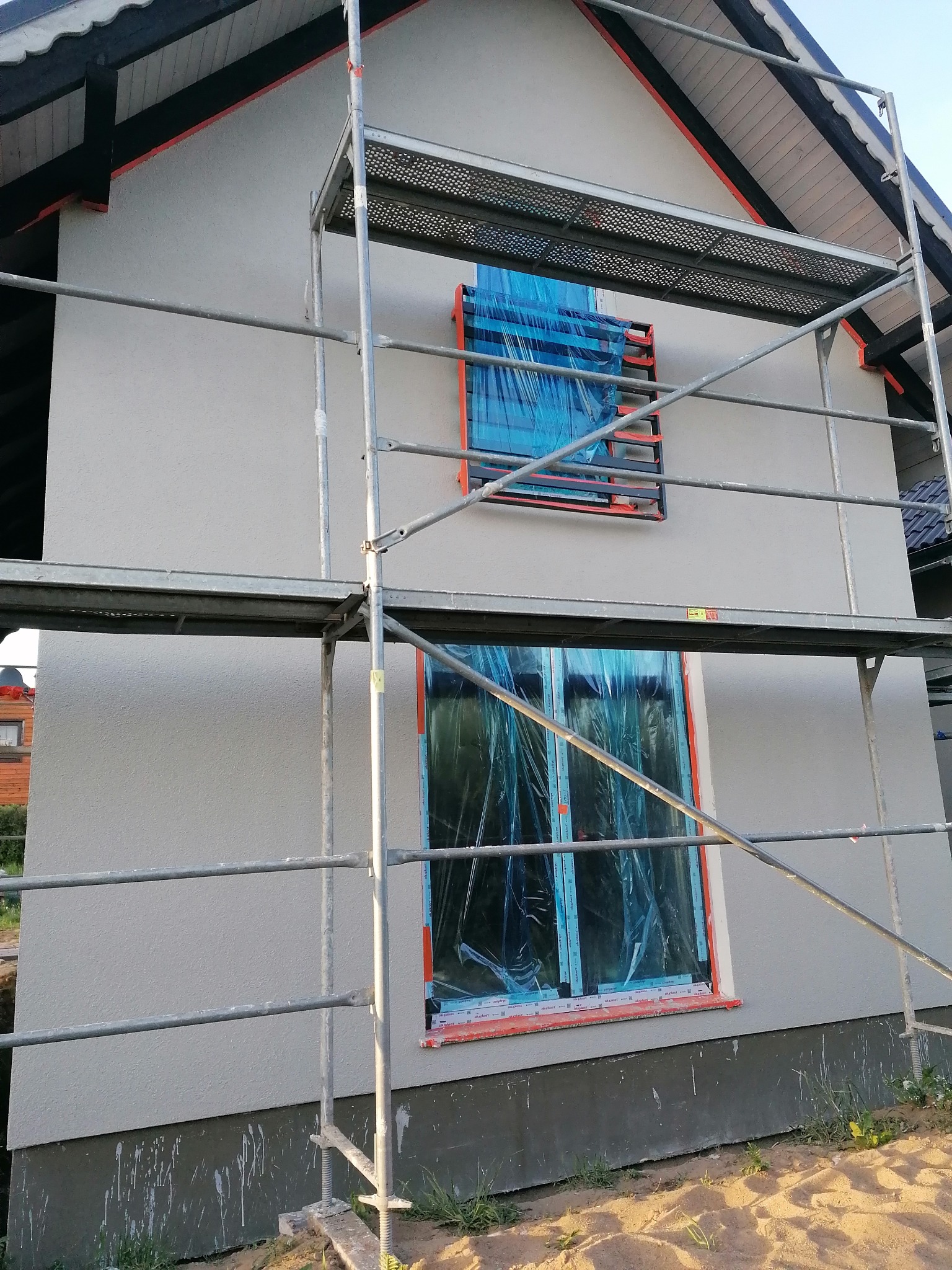 Dom w trakcie nakładania jasnoszarego tynku elewacyjnego, okna zabezpieczone folią, widoczne rusztowanie i fragment podmurówki z zaciekami.