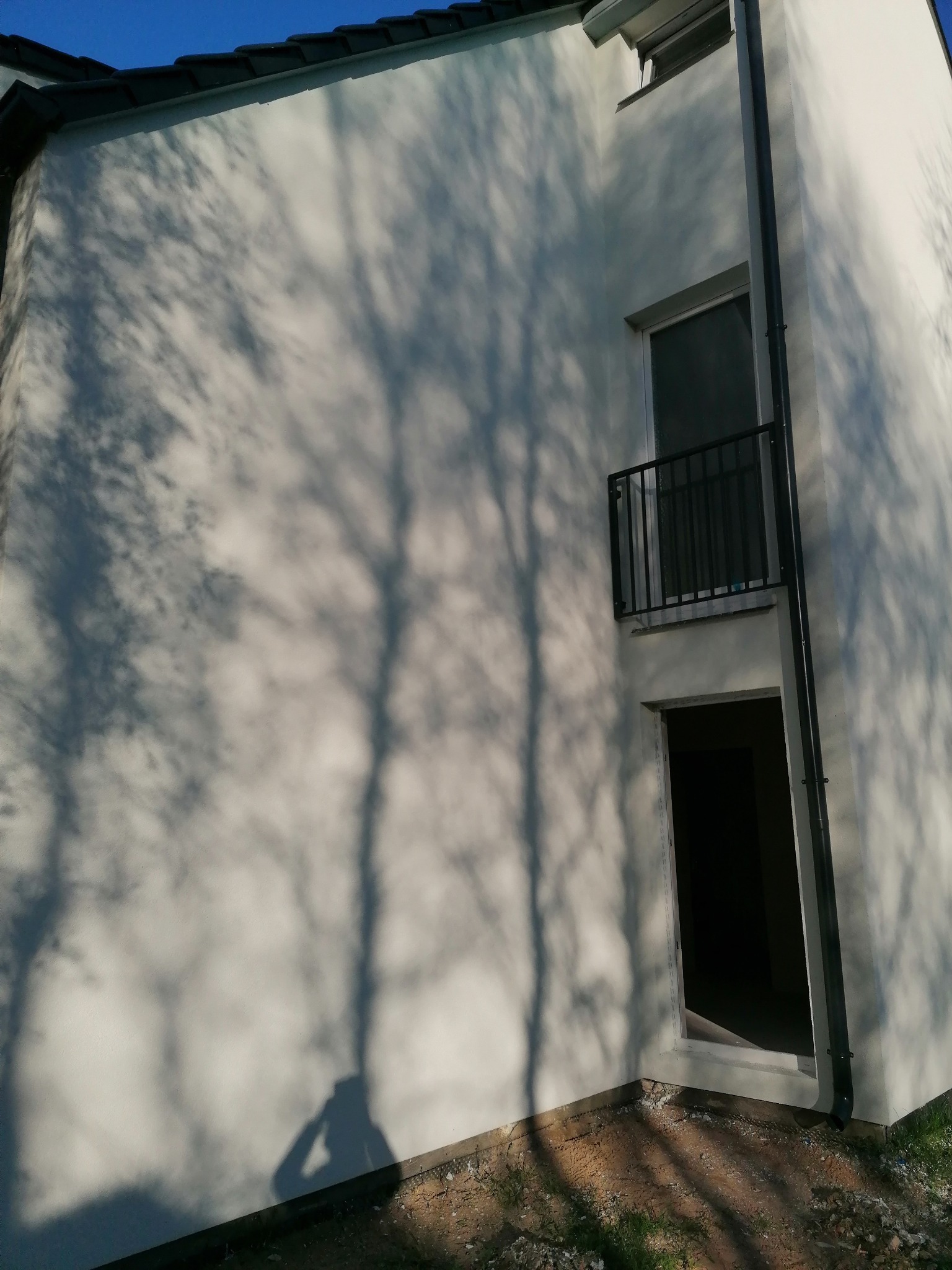 Świeżo otynkowana elewacja budynku z widocznymi cieniami drzew, balkonem z czarną balustradą i rynną, w słoneczny dzień.