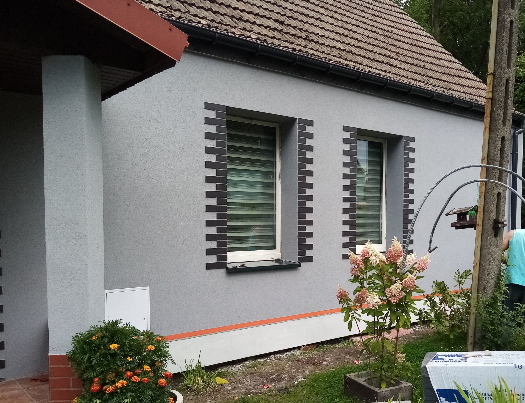 Szara elewacja domu jednorodzinnego z dekoracyjnym obramowaniem okien z ciemnej cegły klinkierowej, widoczne kwiaty w donicach i krzewach przed budynkiem.