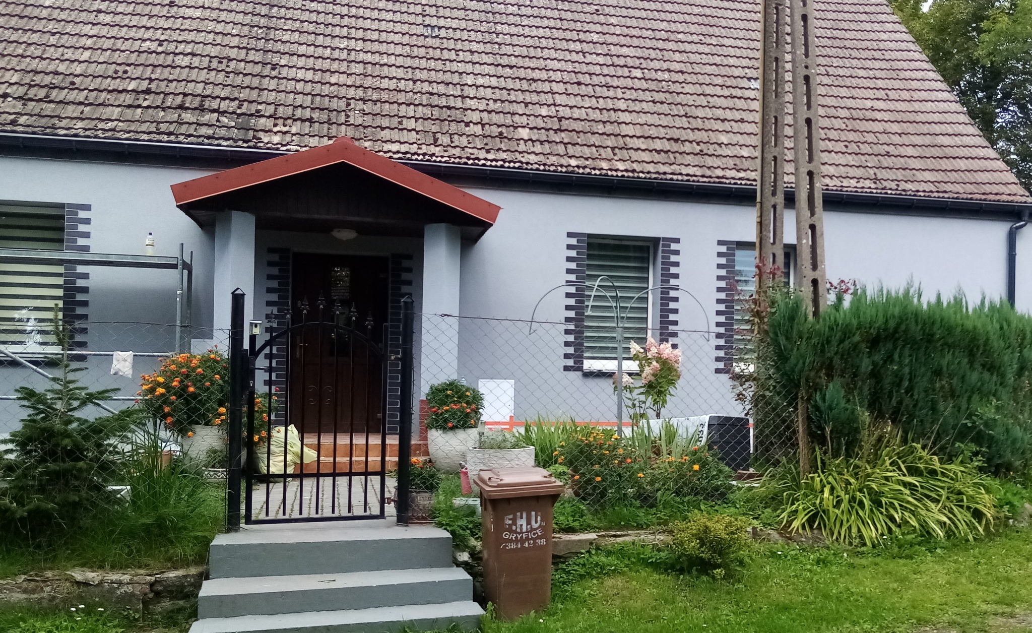 Szary dom jednorodzinny z brązowym dachem, czarną bramą i elementami dekoracyjnymi z cegły wokół okien, częściowo zasłonięty rusztowaniem i bujną roślinnością w ogrodzie.