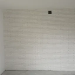 Ściana wykończona białą cegłą dekoracyjną, z widocznymi puszkami elektrycznymi i otworem wentylacyjnym, w trakcie prac wykończeniowych.