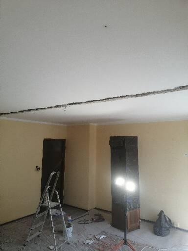 Pokój w trakcie remontu, widoczna drabina, narzędzia, podłoga zabezpieczona folią, ściany w jasnym kolorze, sufit z widocznym uszkodzeniem na styku ze ścianą, oświetlenie halogenowe.