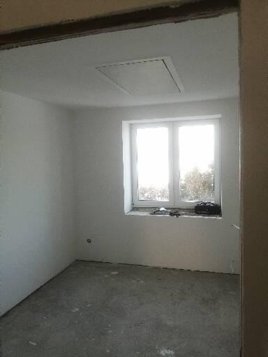 Pusty pokój w trakcie remontu, ściany i sufit pokryte świeżym tynkiem, okno z widokiem na zewnątrz, na parapecie leżą narzędzia.