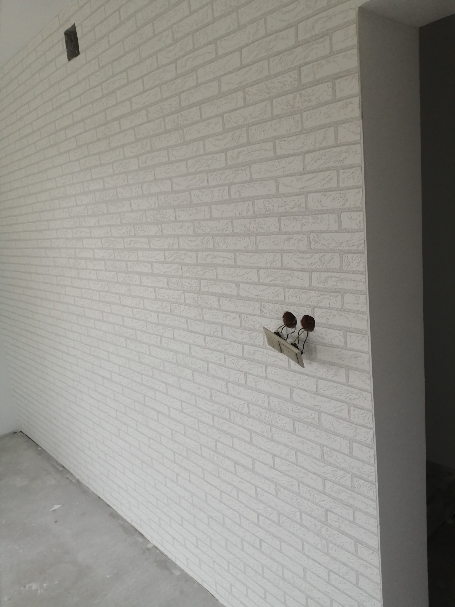 Ściana wykończona tapetą imitującą białą cegłę, z wystającymi przewodami elektrycznymi i puszką instalacyjną, surowy beton na podłodze.