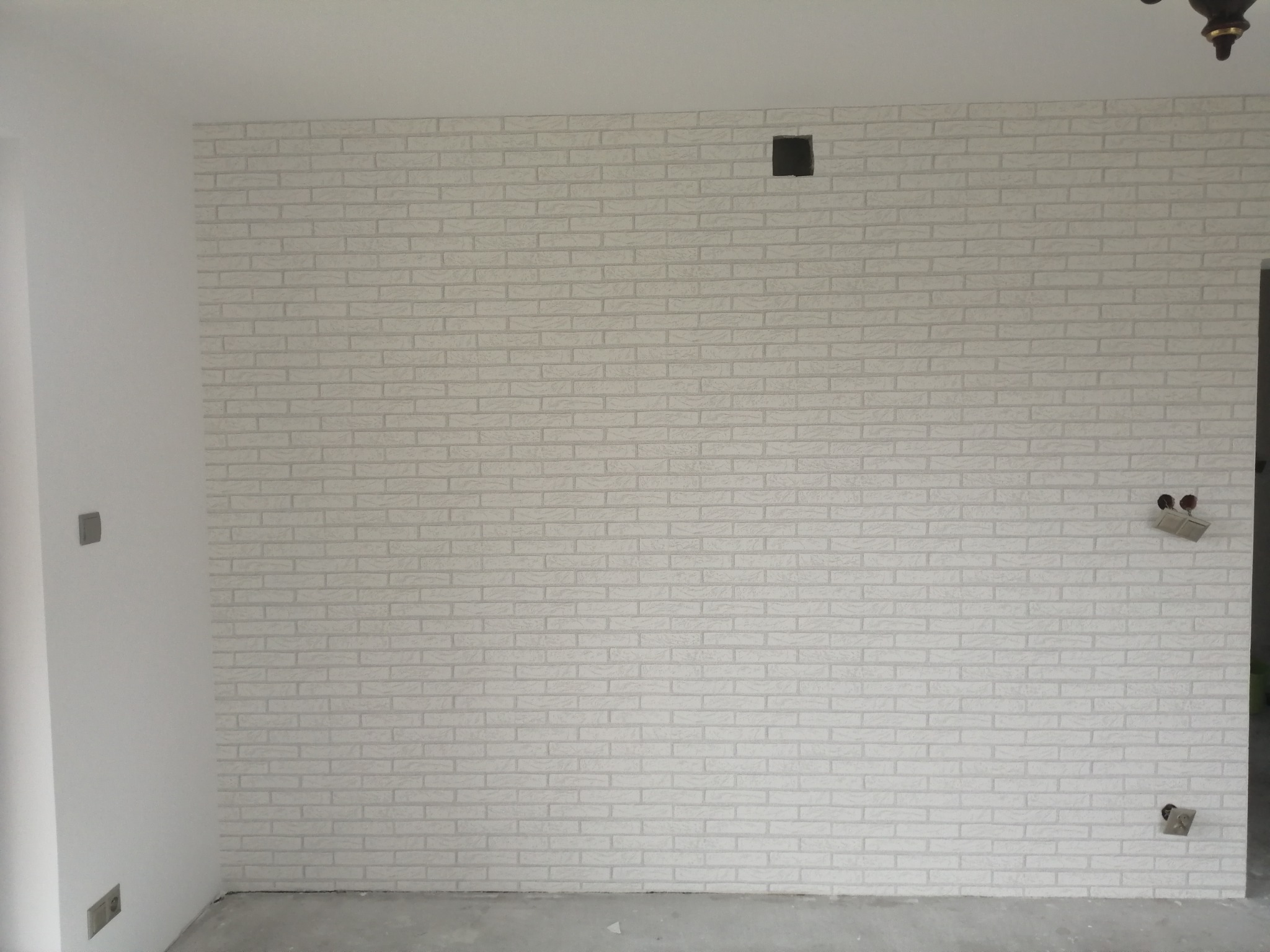 Ściana wykończona białą cegłą dekoracyjną, z widocznymi puszkami elektrycznymi i otworem wentylacyjnym, w trakcie prac wykończeniowych.