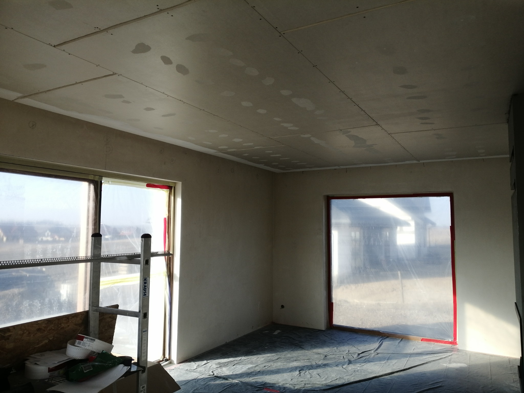 Pokój w trakcie remontu: sufit i ściany pokryte płytami kartonowo-gipsowymi, widoczne ślady szpachlowania, okna zabezpieczone folią, drabina Krause.