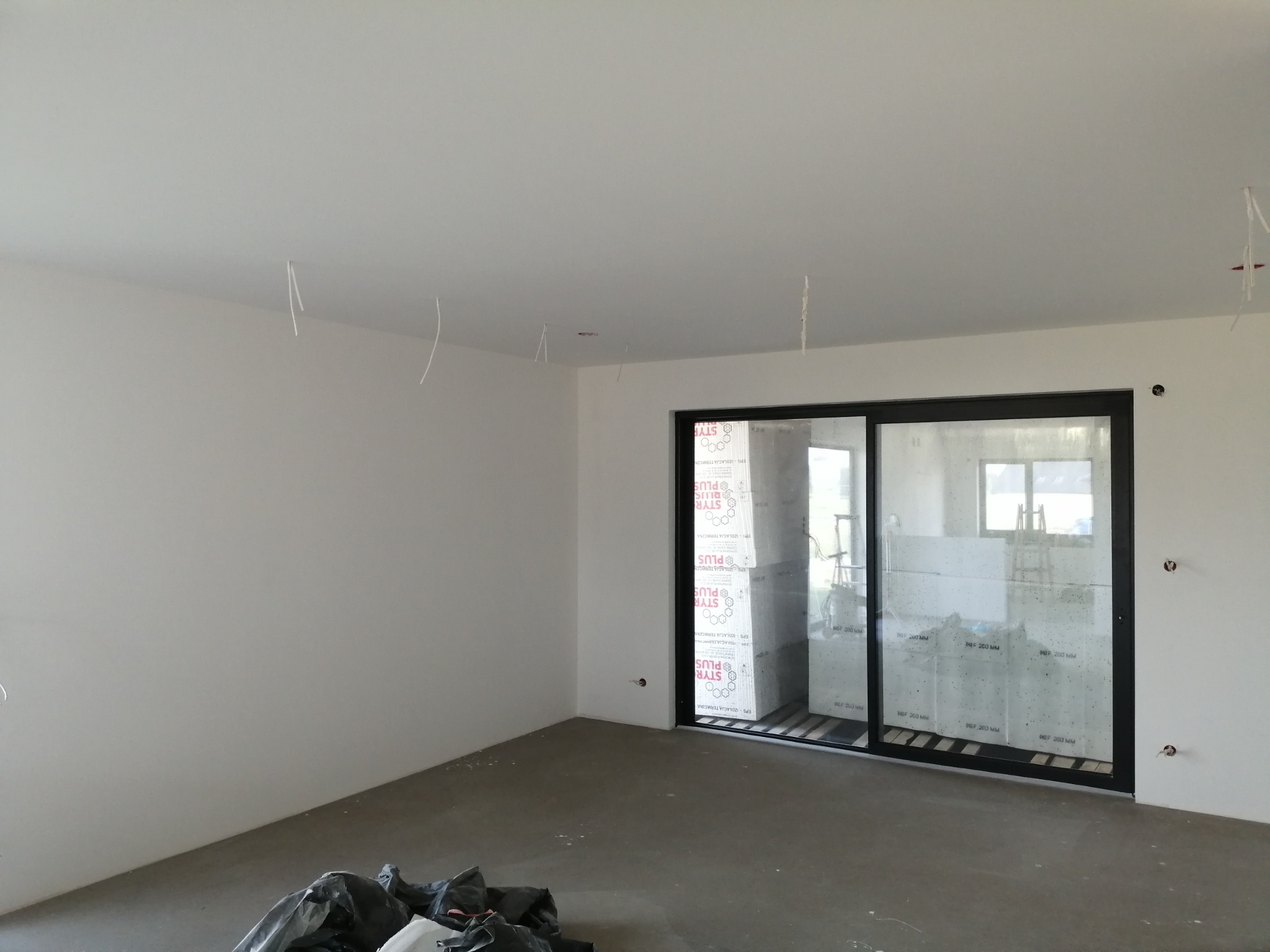 Pokój w trakcie remontu, ściany świeżo pomalowane na biało, widoczne wystające kable z sufitu, duże okno przesuwne z widokiem na styropian izolacyjny.