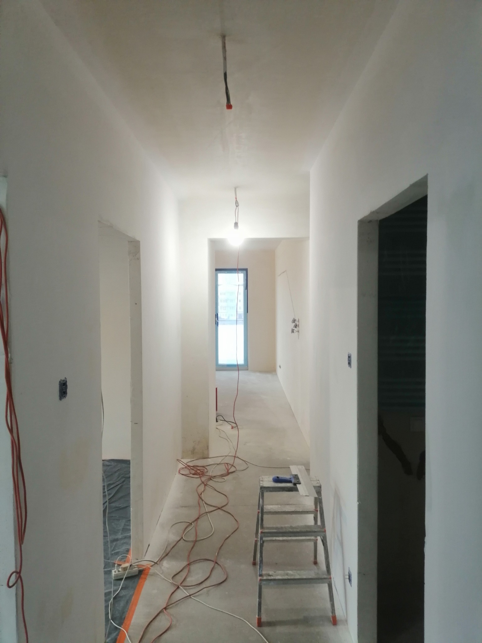 Korytarz w trakcie remontu z świeżo wygładzonymi ścianami, aluminiowa drabina z narzędziami, widoczne kable elektryczne i zabezpieczona podłoga.