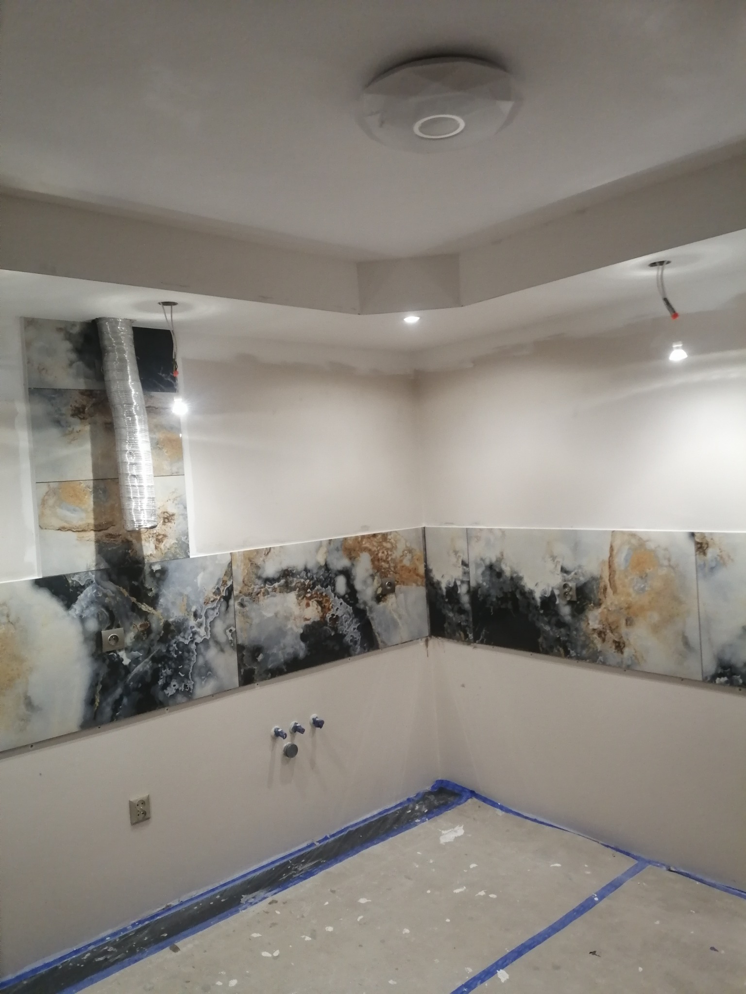 Fragment wnętrza w trakcie prac wykończeniowych: ściany częściowo pokryte płytkami imitującymi marmur, zabezpieczona podłoga, widoczne instalacje elektryczne i hydrauliczne, sufit z oświetleniem...