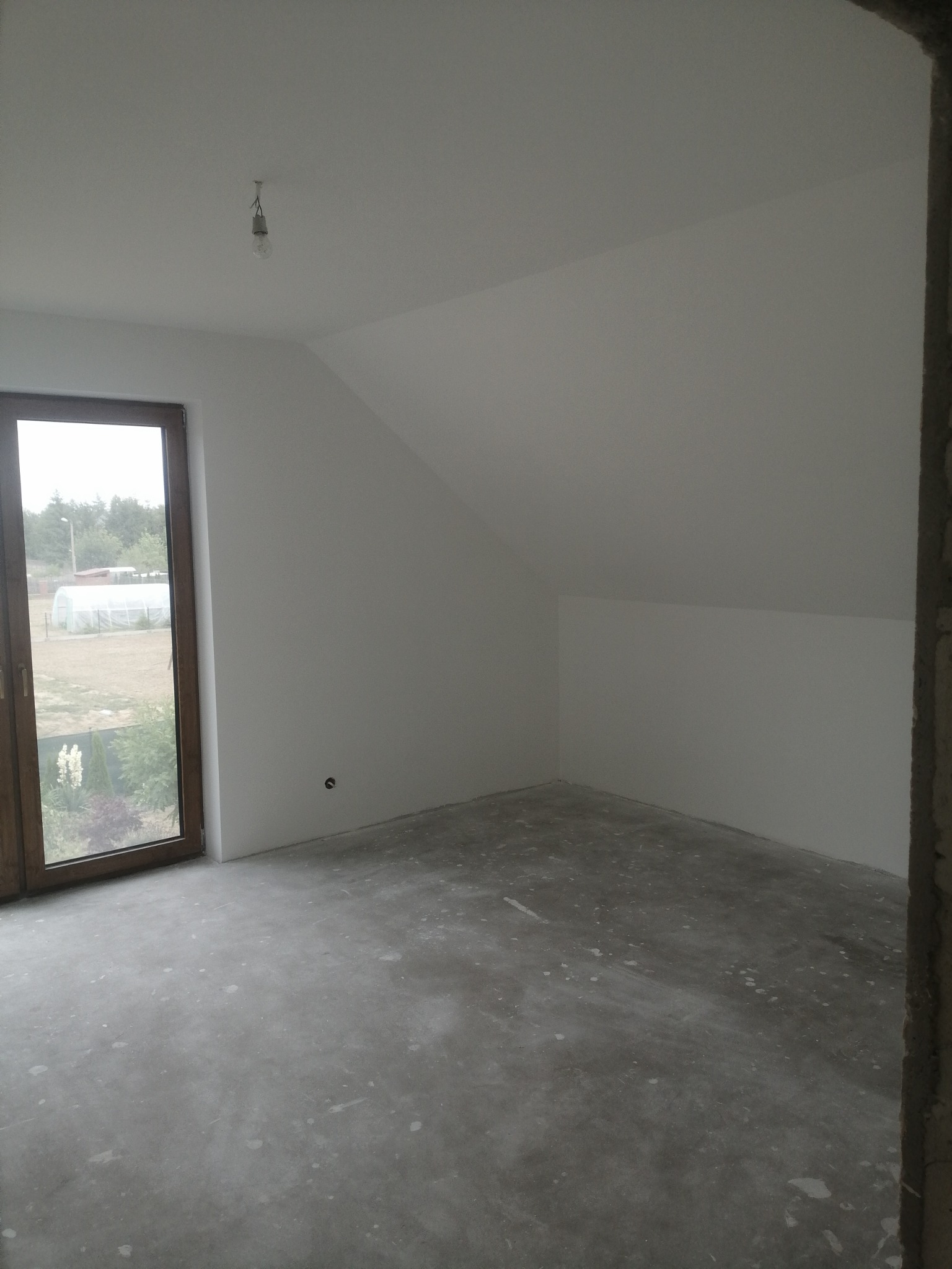 Pokój na poddaszu w stanie deweloperskim z oknem i surową żarówką, ściany pomalowane na biało, podłoga betonowa z widocznymi śladami po pracach budowlanych.