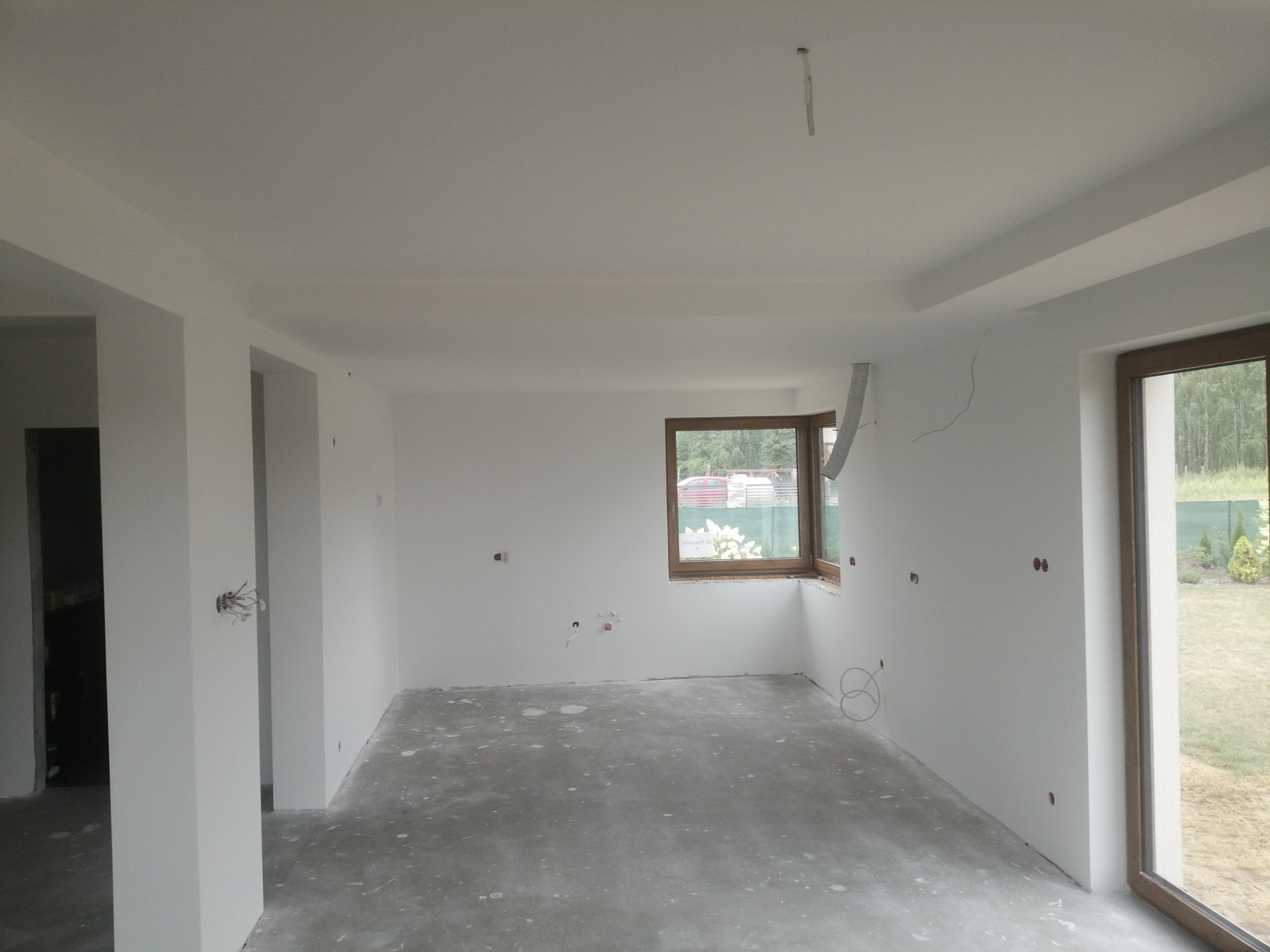 Surowe wnętrze w trakcie remontu, świeżo pomalowane na biało ściany i sufit, widoczne wystające kable elektryczne i instalacje, betonowa podłoga z plamami, okna z drewnianymi ramami.