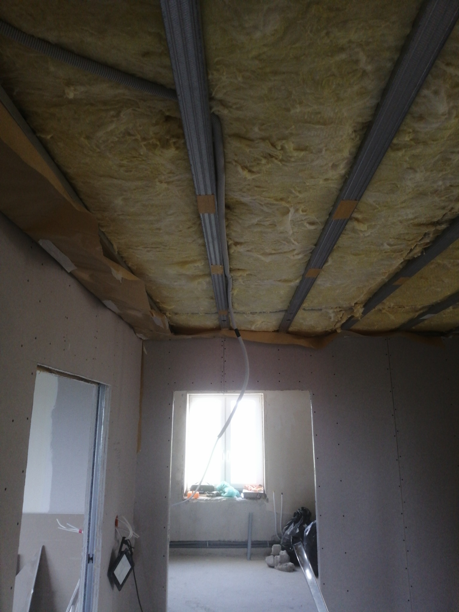 Montaż sufitu podwieszanego: widoczne profile metalowe, izolacja z wełny mineralnej oraz płyty kartonowo-gipsowe przed szpachlowaniem, okno w tle.