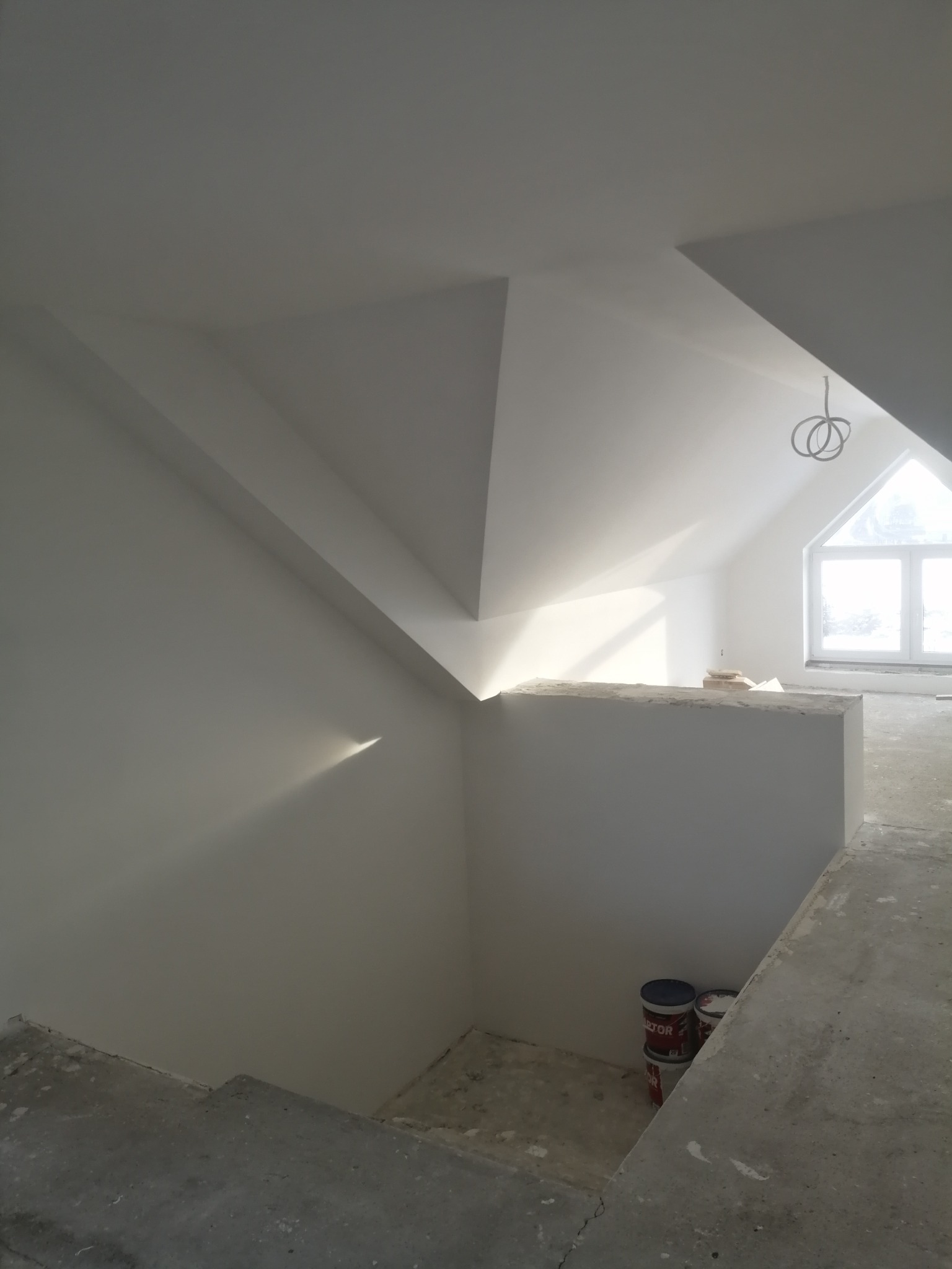 Poddasze w trakcie prac wykończeniowych, widoczne gładkie, białe ściany, okno dachowe, surowa podłoga i puszki z farbą w rogu.