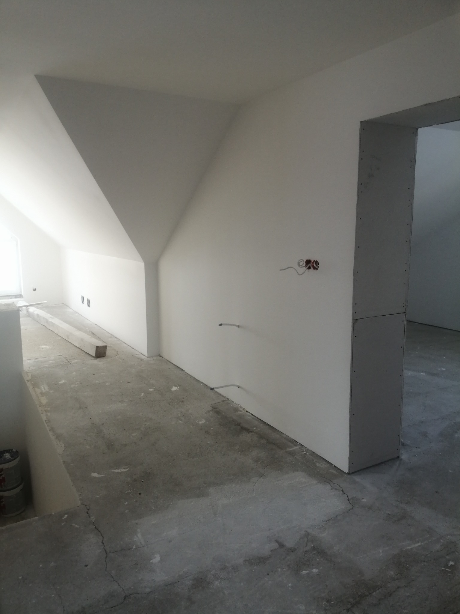 Poddasze w trakcie prac wykończeniowych, widoczne ściany z płyt kartonowo-gipsowych, surowy betonowy podkład, przygotowane otwory na gniazdka elektryczne i listwa przypodłogowa.