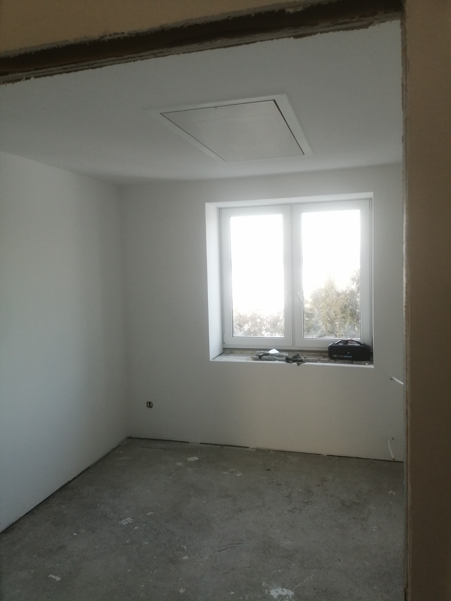 Pusty pokój po remoncie, ściany pomalowane na biało, surowy beton na podłodze, widoczny otwór rewizyjny w suficie, okno z widokiem na drzewa, radio budowlane na parapecie.