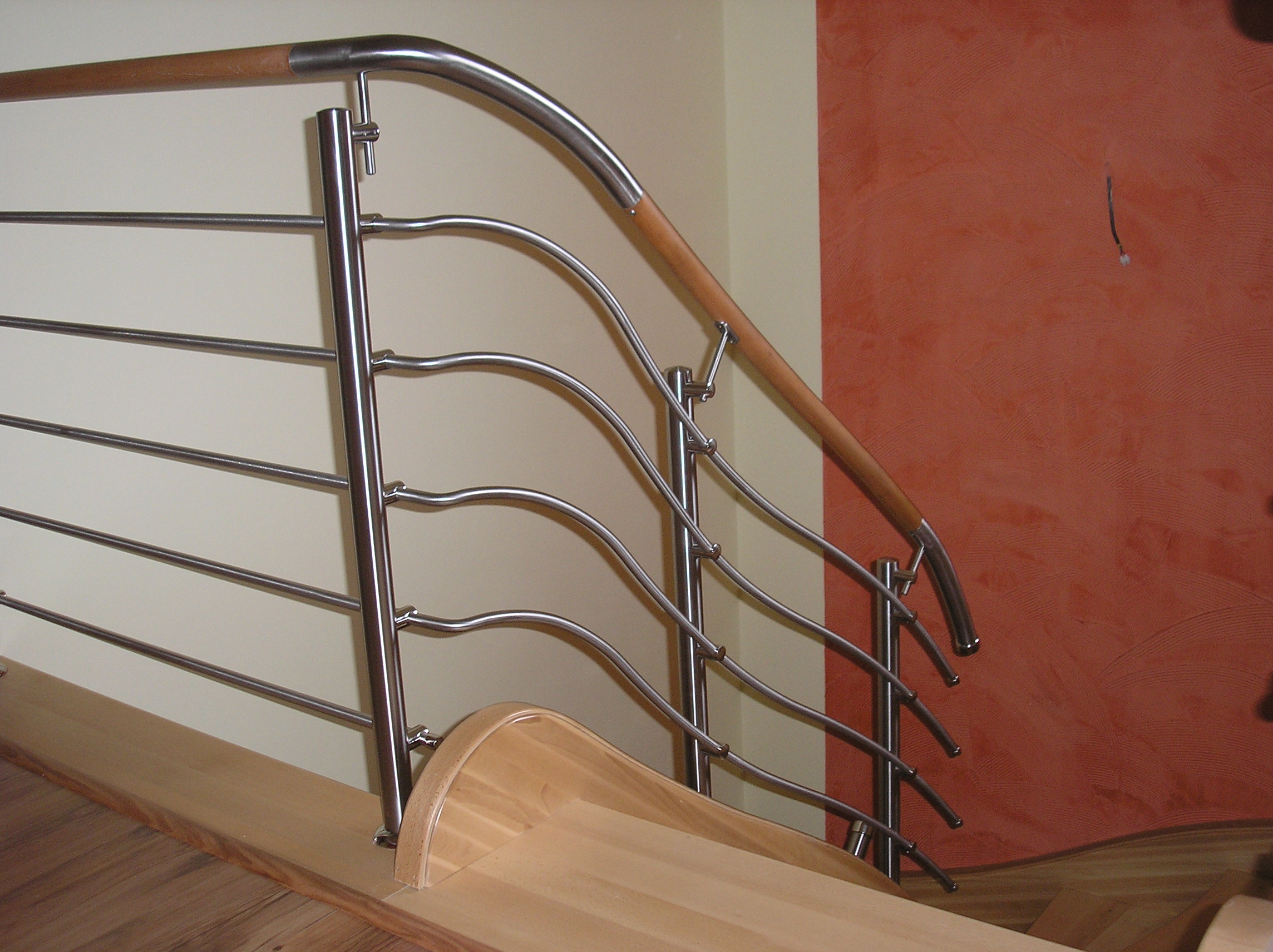 Nowoczesna balustrada schodowa ze stali nierdzewnej z drewnianym pochwytem, przymocowana do drewnianych stopni schodów, na tle ściany w odcieniach beżu i terakoty.