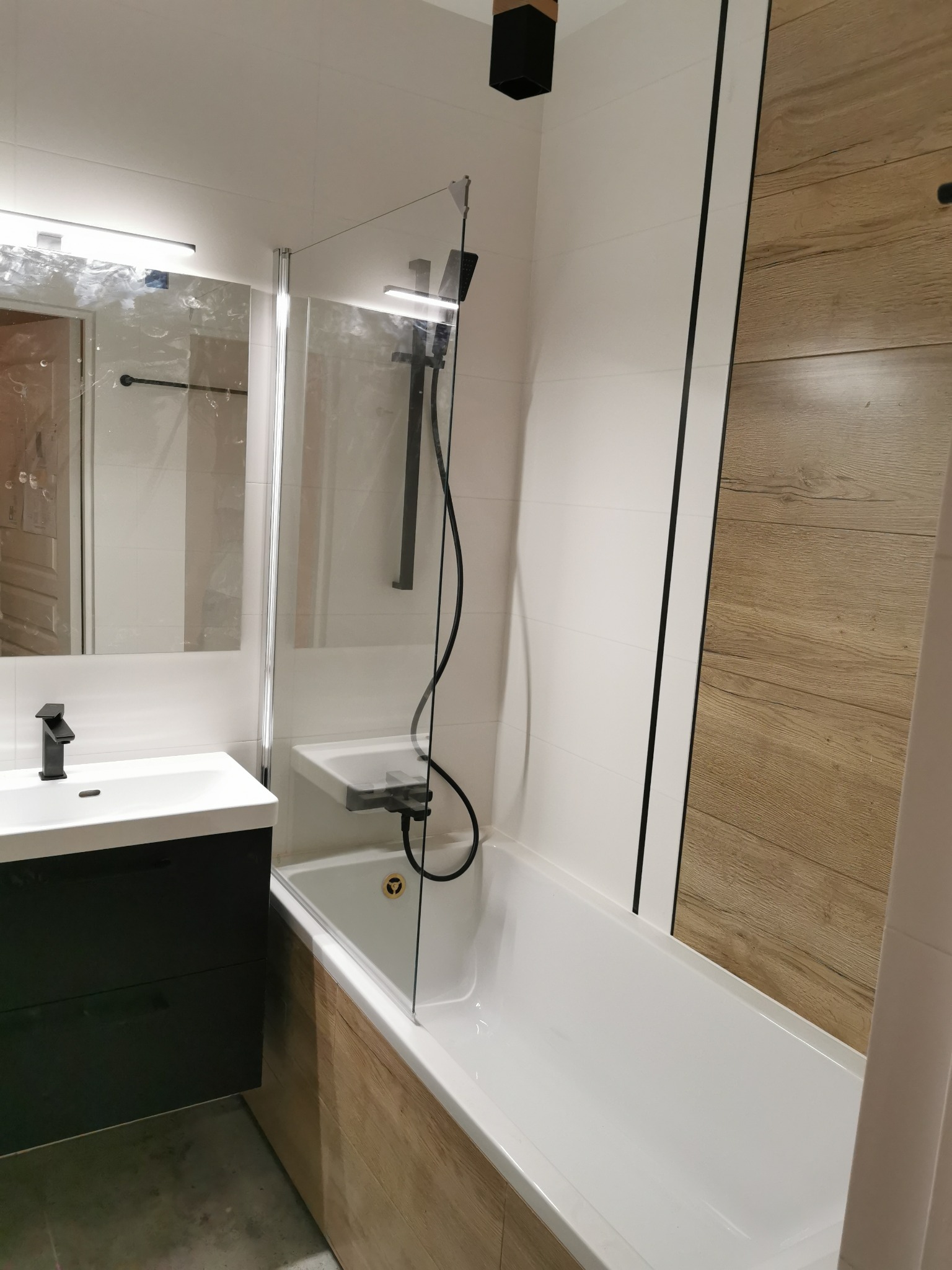 Nowoczesna łazienka z białą wanną obudowaną drewnopodobnymi panelami, czarną armaturą, szklanym parawanem nawannowym i umywalką z szafką w kolorze grafitowym.