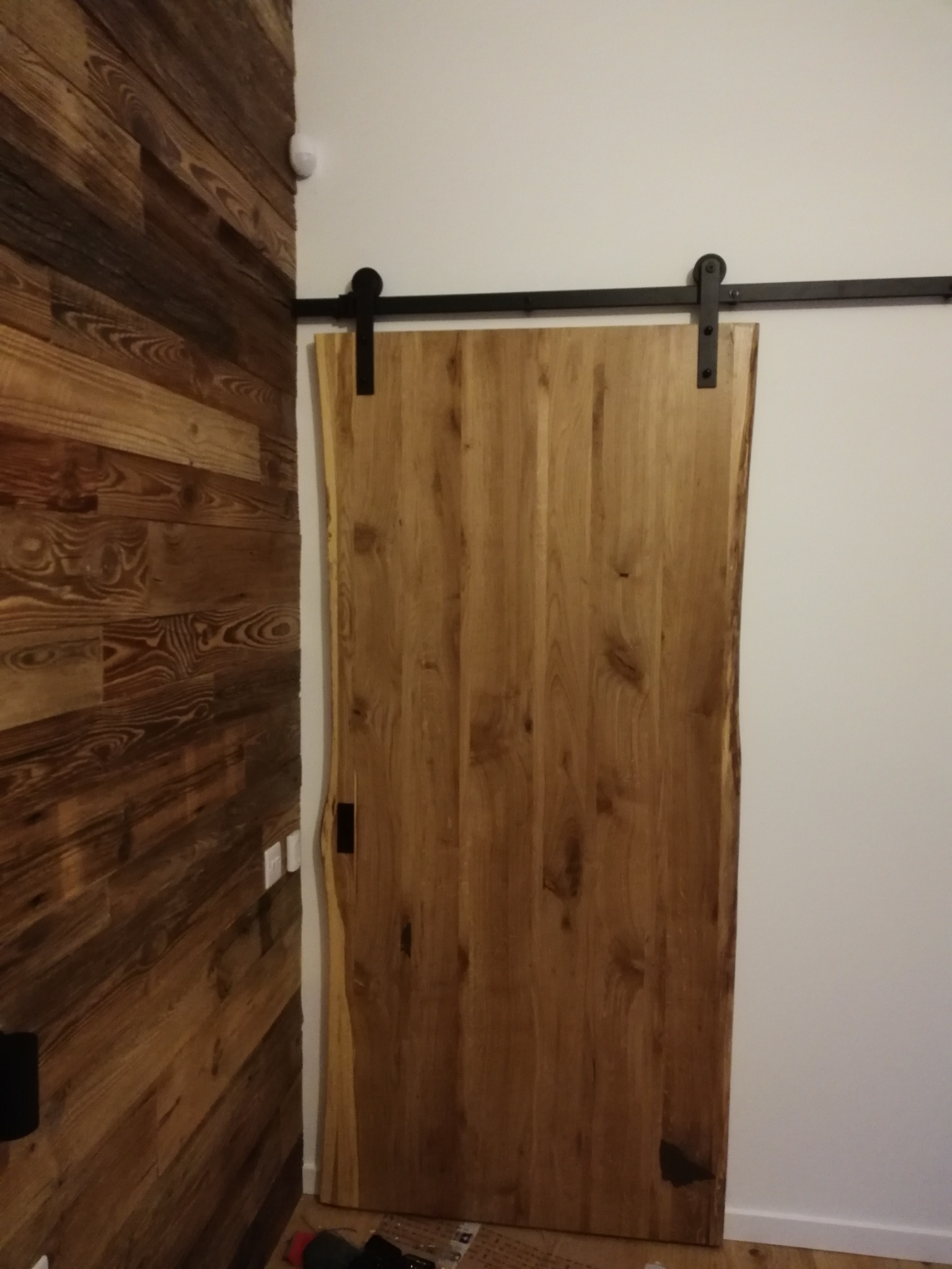 Surowe drewniane drzwi przesuwne na czarnej szynie, z naturalną krawędzią, na tle ściany z desek o bogatym usłojeniu i białej ściany.