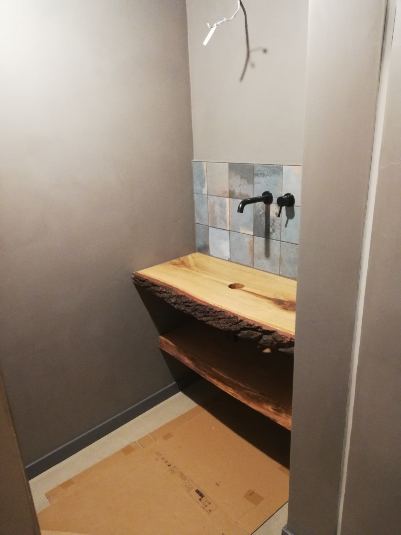 Łazienka w trakcie wykańczania: drewniany blat umywalki z naturalną krawędzią, czarna bateria ścienna na tle mozaiki, podłoga zabezpieczona kartonem, widoczne wystające kable.