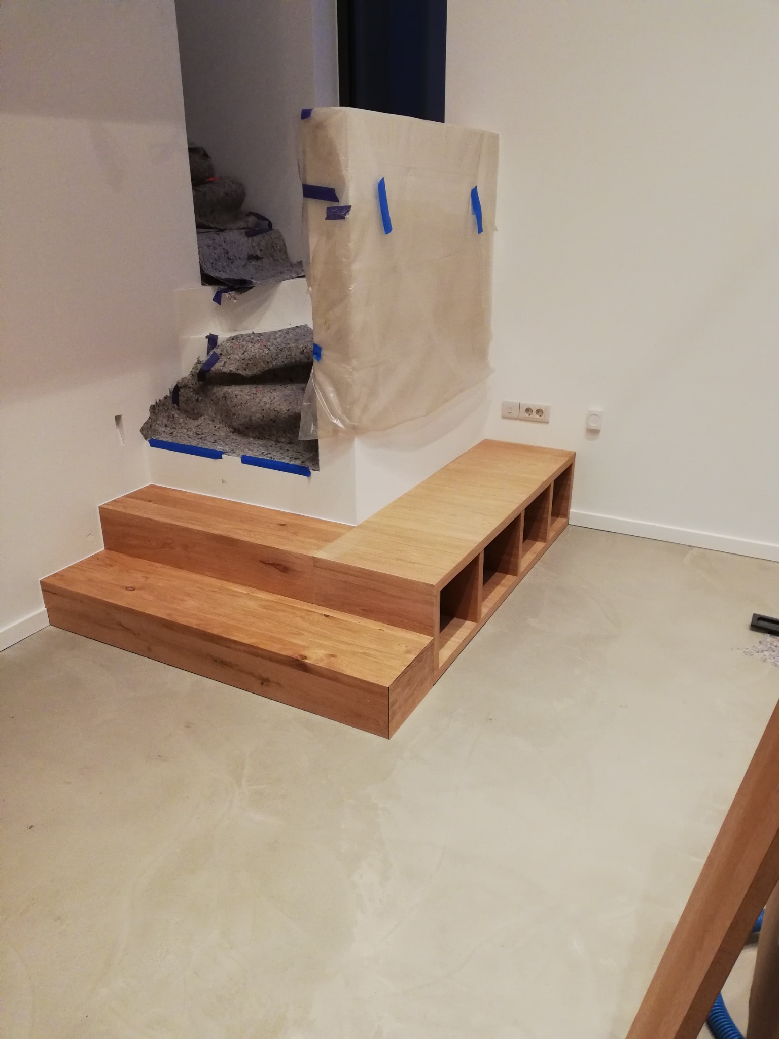 Drewniana konstrukcja schodkowa z wbudowanymi półkami, wykonana z jasnego drewna, umieszczona w surowym wnętrzu podczas prac wykończeniowych, z widocznymi zabezpieczeniami i materiałami budowlanymi...