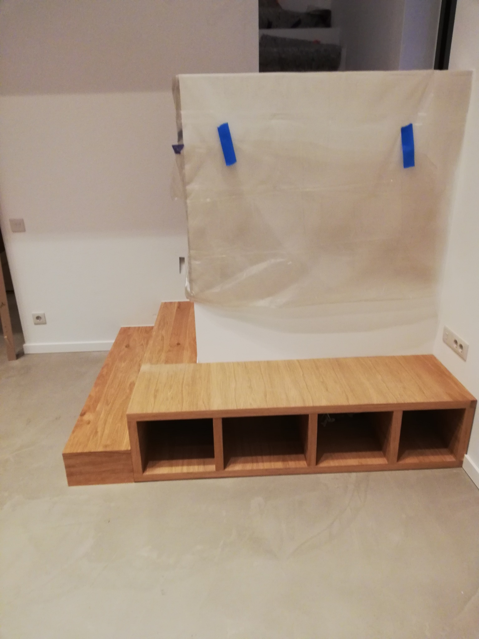 Drewniana konstrukcja antresoli w trakcie budowy, zabezpieczona folią malarską, z widocznymi schodkami i szafką z przegródkami, na tle białych ścian.