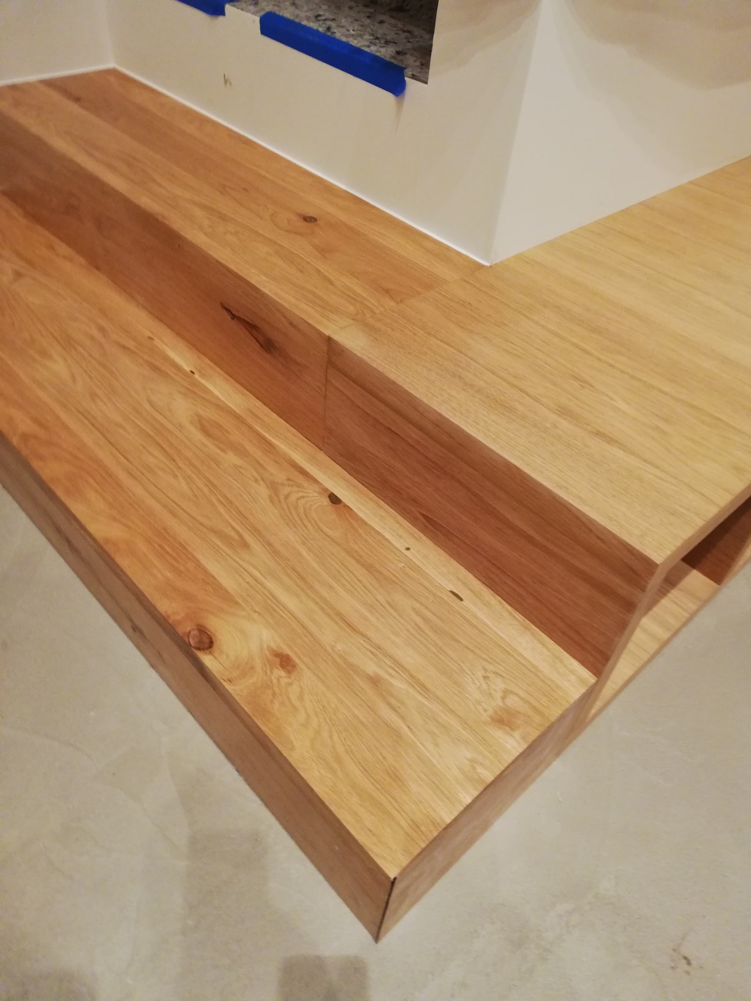 Narożny element wykończeniowy z jasnego drewna, prawdopodobnie schodek lub podest, z widocznymi usłojeniami i sękami, z fragmentem ściany zabezpieczonej niebieską taśmą malarską w tle.