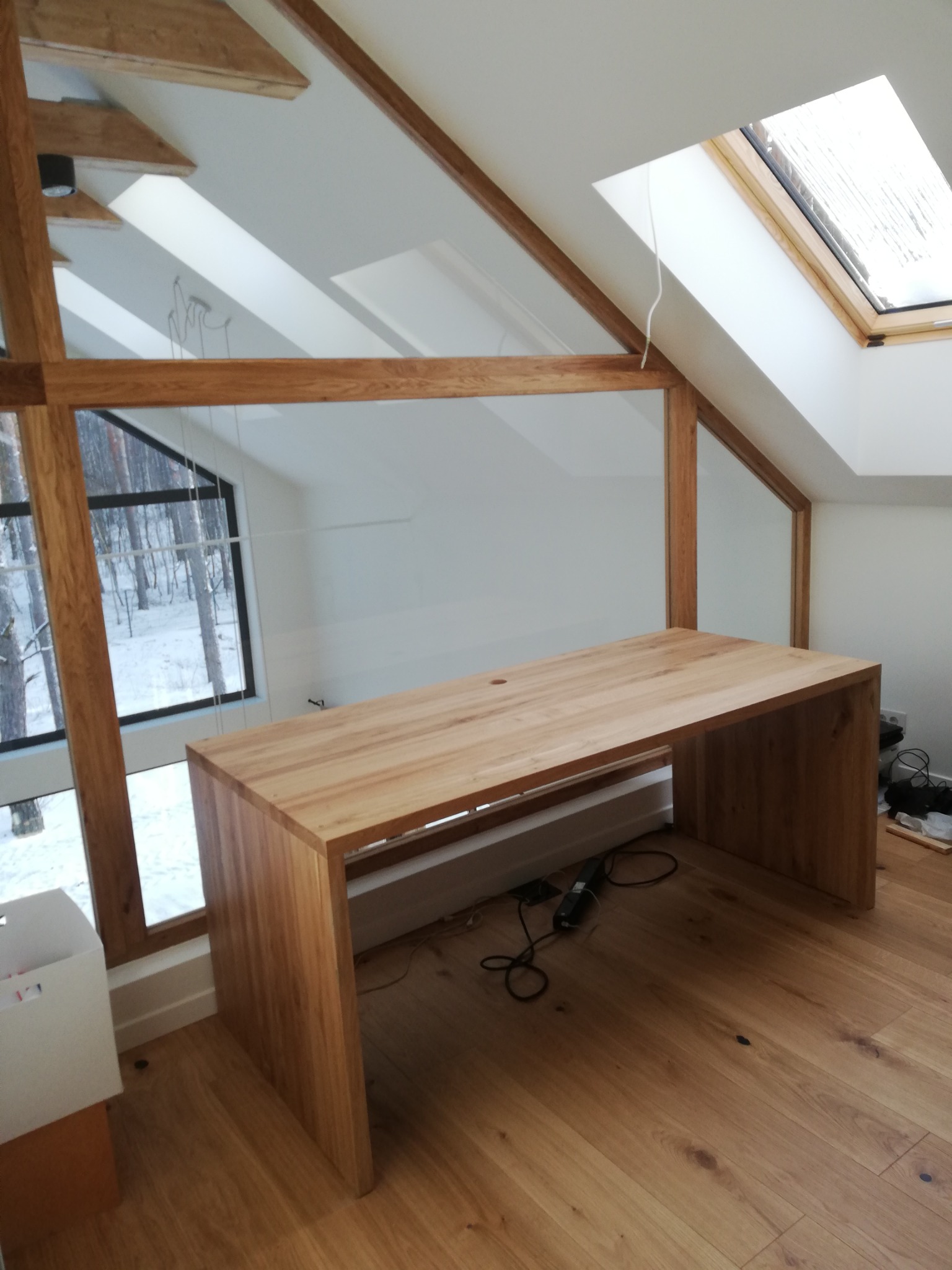 Drewniany biurko w jasnym pokoju na poddaszu z oknem dachowym i dużym oknem z widokiem na zasypany śniegiem las. Biurko wykonane z jasnego drewna, stojące na drewnianej podłodze. Widoczne przewody...